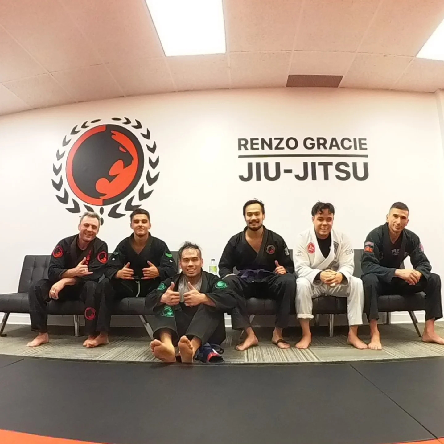 Seis praticantes de kimono sentados sob o logo da academia depois da aula