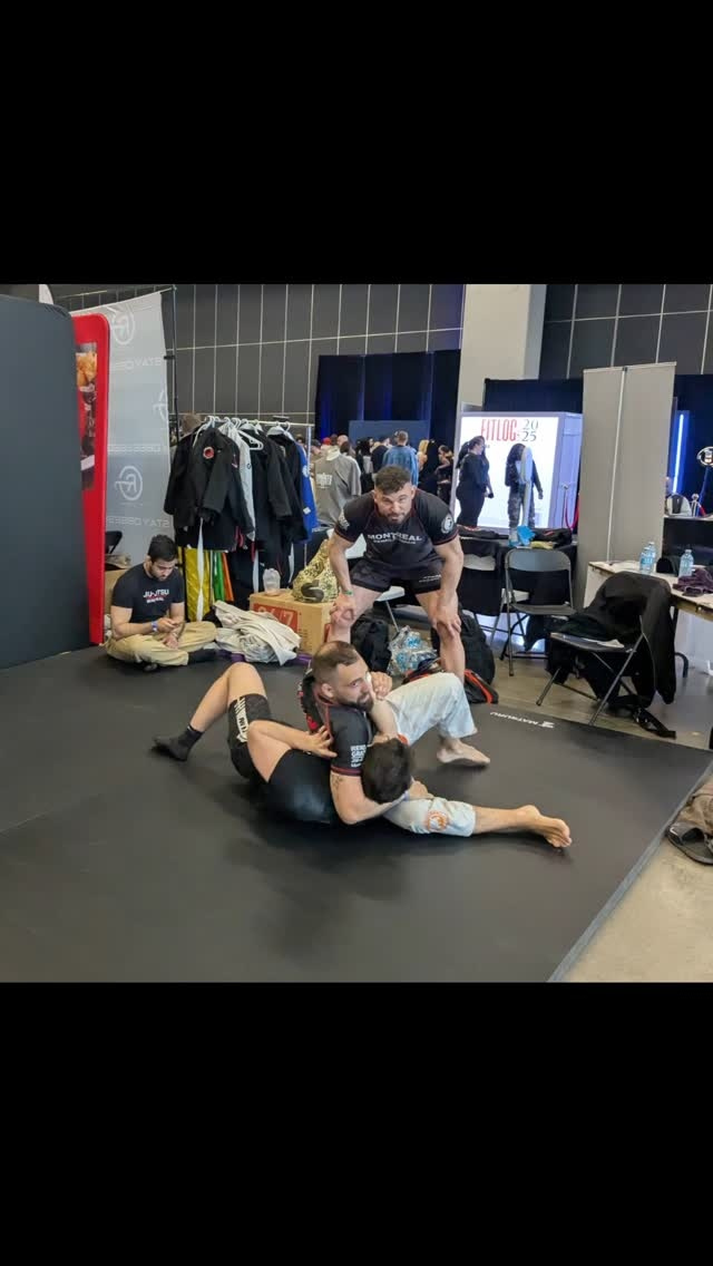 A grappling demonstration on black mats at an event booth, with gear on racks behind