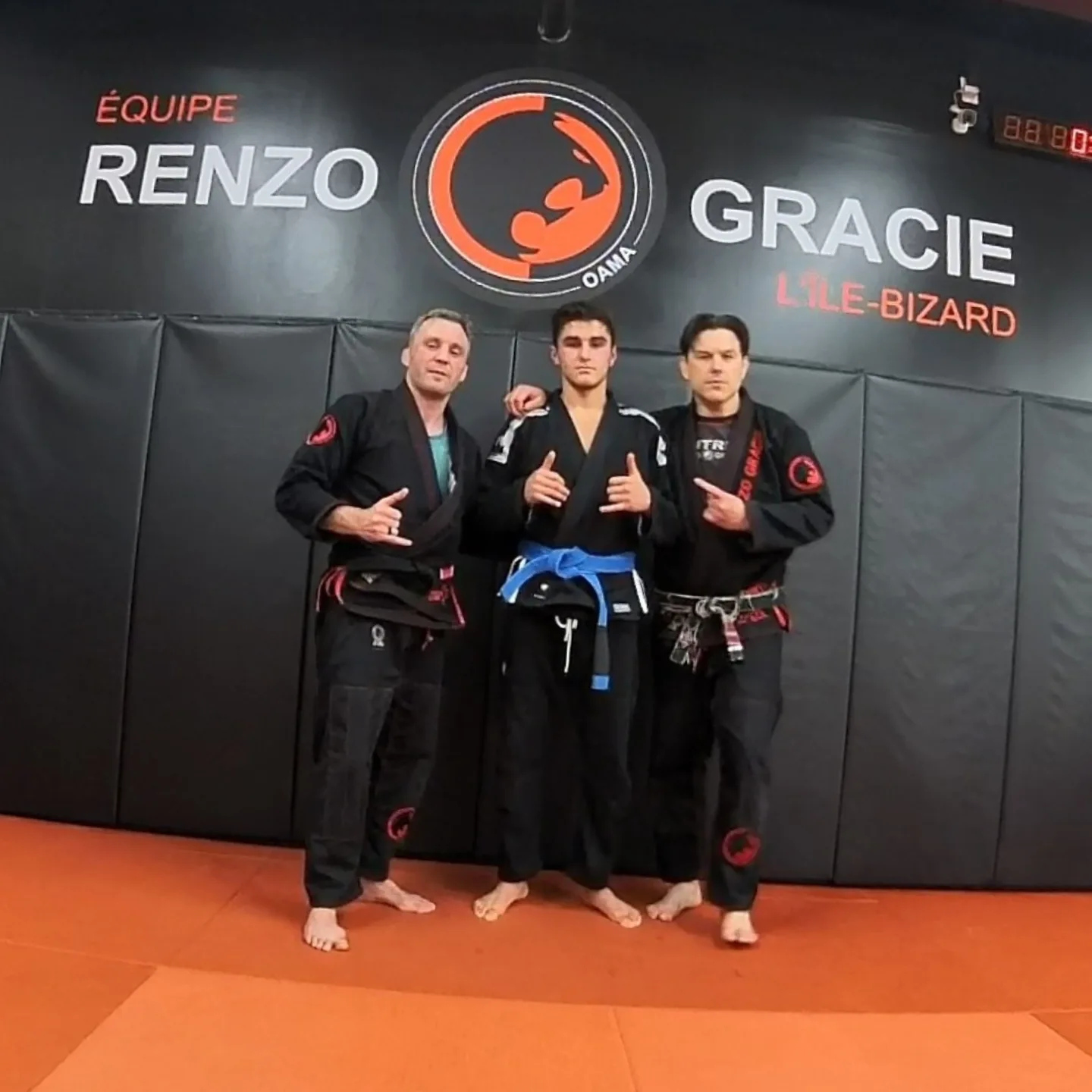 A young blue belt competitor stands between two coaches in black gis in front of a team wall