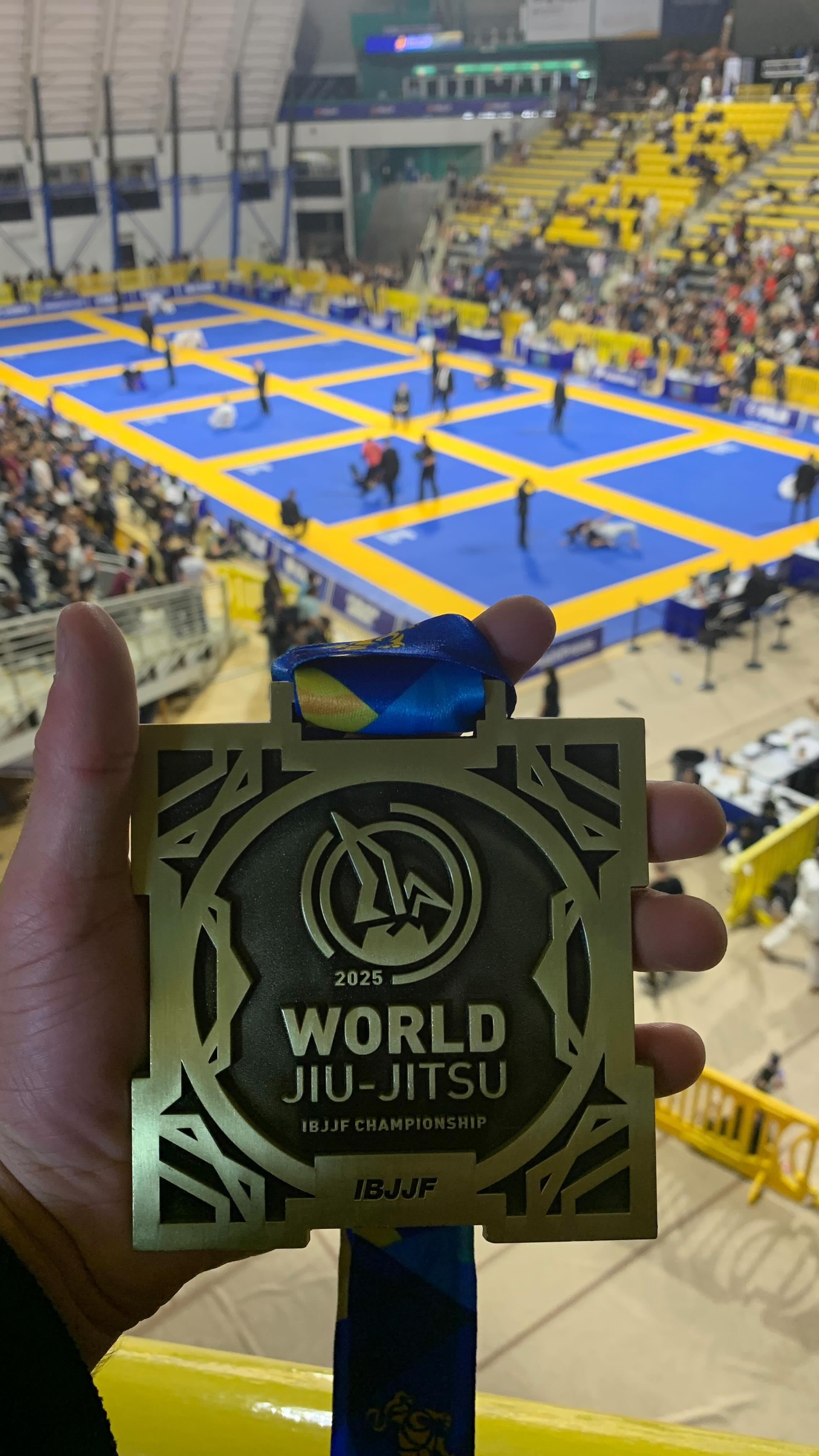 A 2025 World Jiu-Jitsu medal held up above the blue and yellow mats of the arena