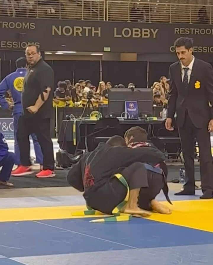 Two competitors in black gis grapple on blue and yellow mats as referees watch