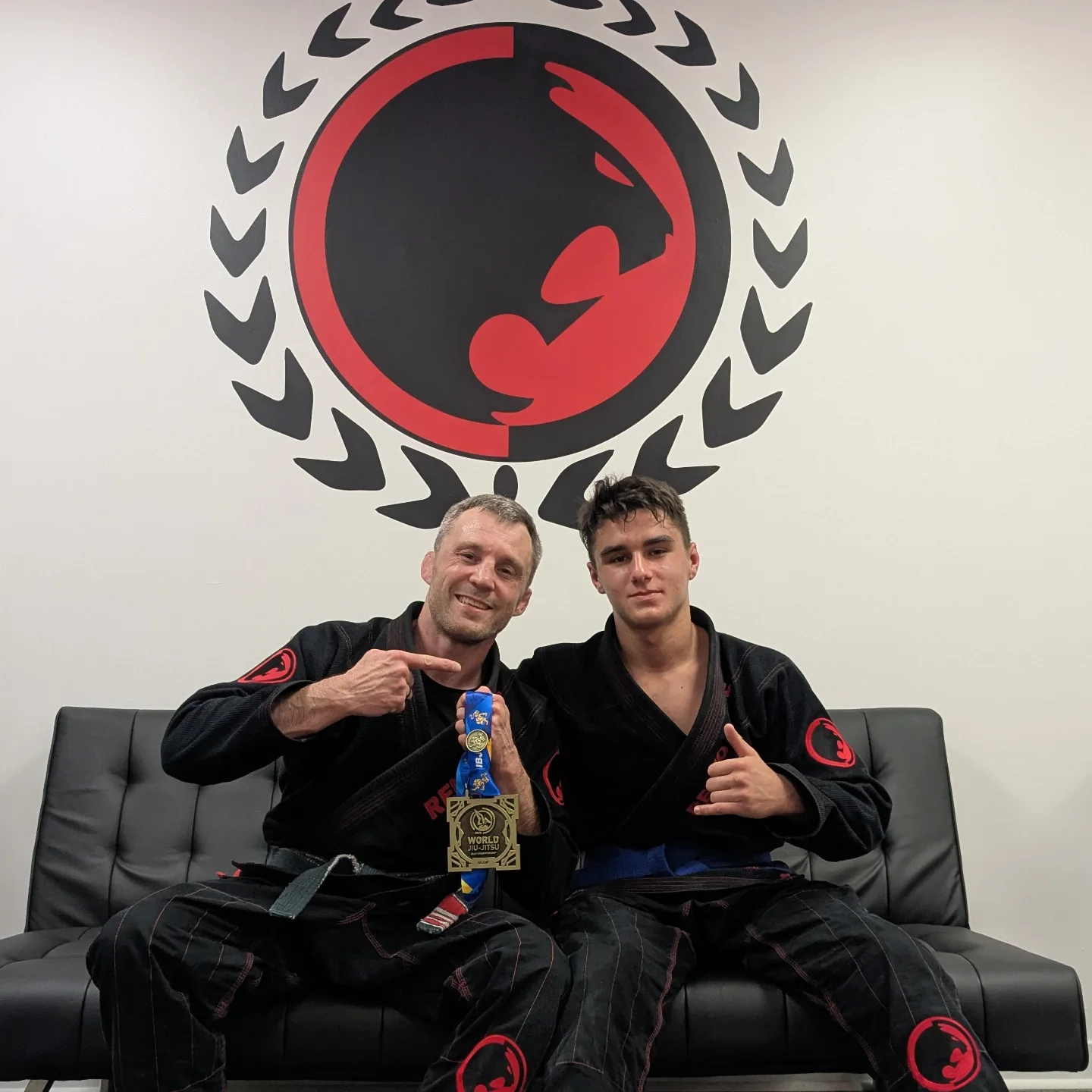 A coach points to the World Jiu-Jitsu medal a young competitor holds up under the academy logo