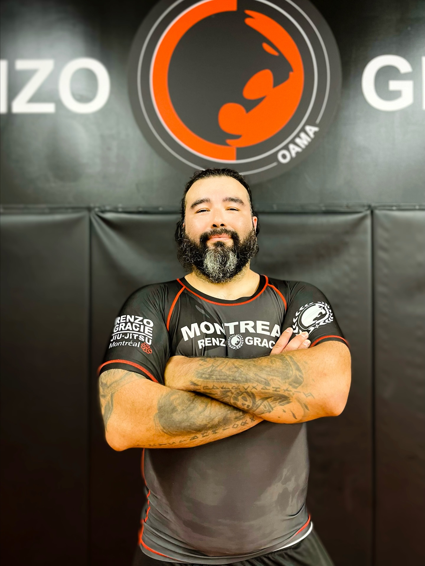 A coach in a team rash guard stands with crossed arms in front of a large red logo