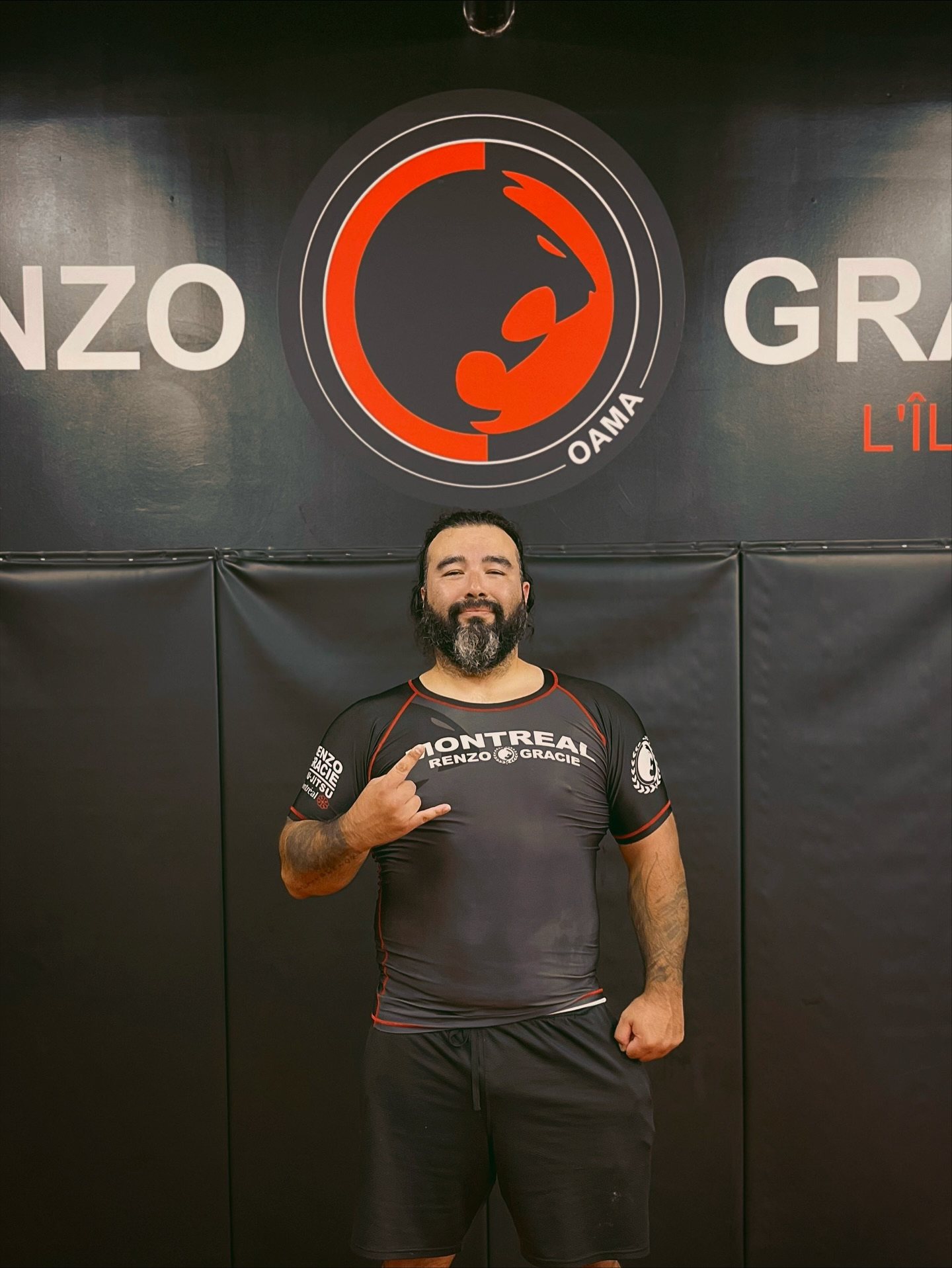 A coach in a team rash guard gives a shaka in front of a large red logo