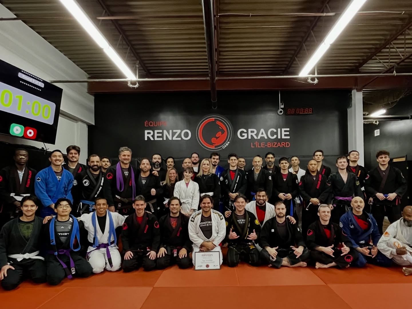 A large group of adults in gis poses on red mats in front of the academy wall