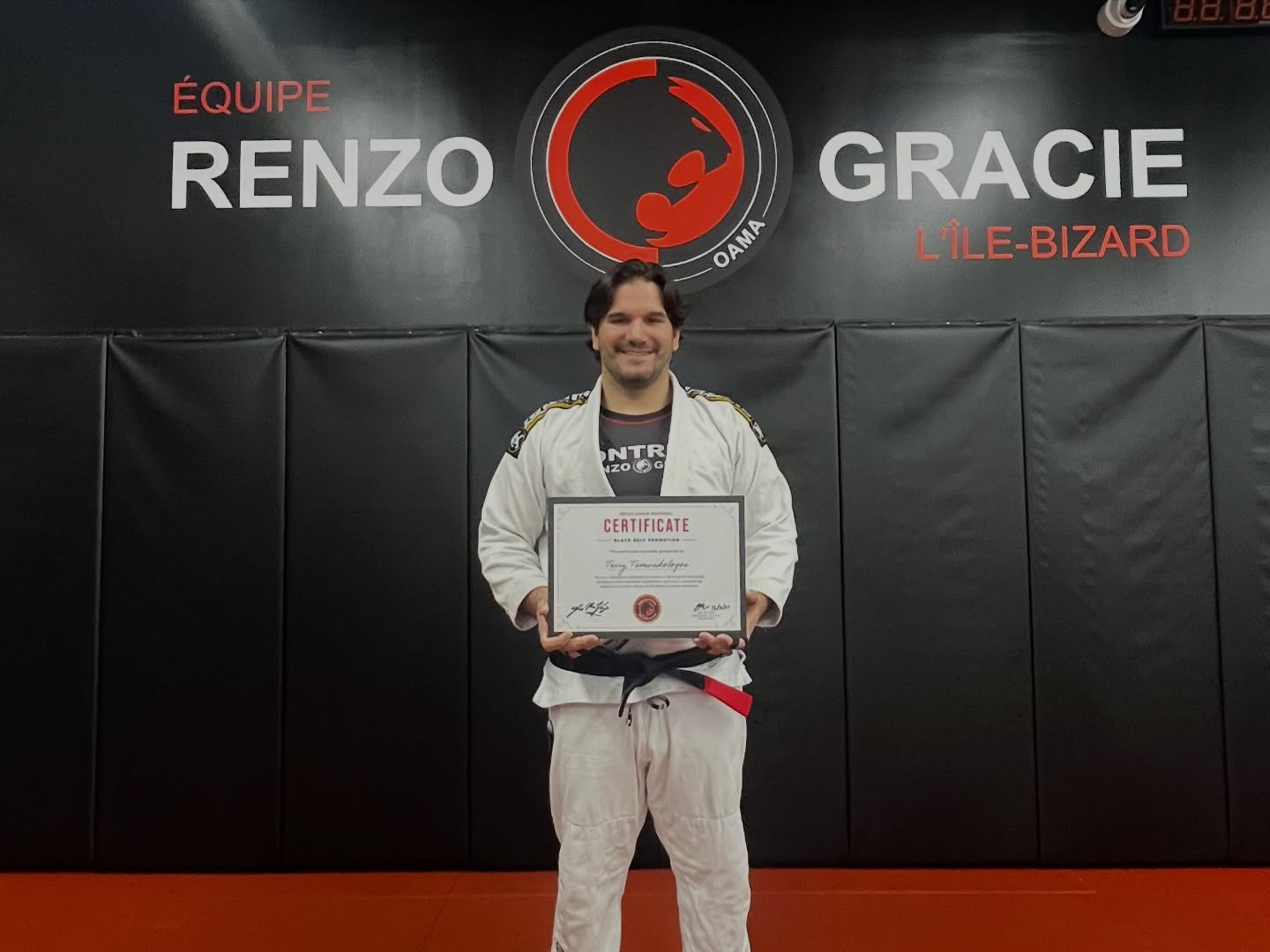 A student in a white gi holds a certificate in front of the academy wall