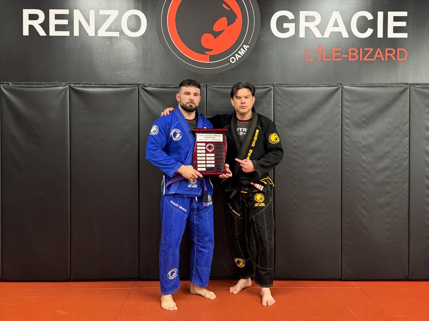 Two people in gis hold an award plaque in front of the academy wall