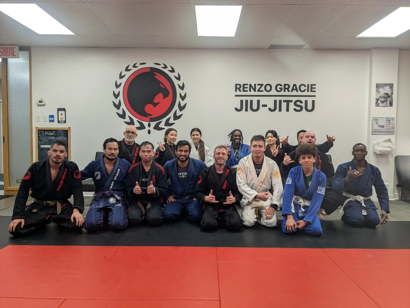 Des adultes en kimono à genoux et debout sous l'enseigne Renzo Gracie Jiu-Jitsu après le cours