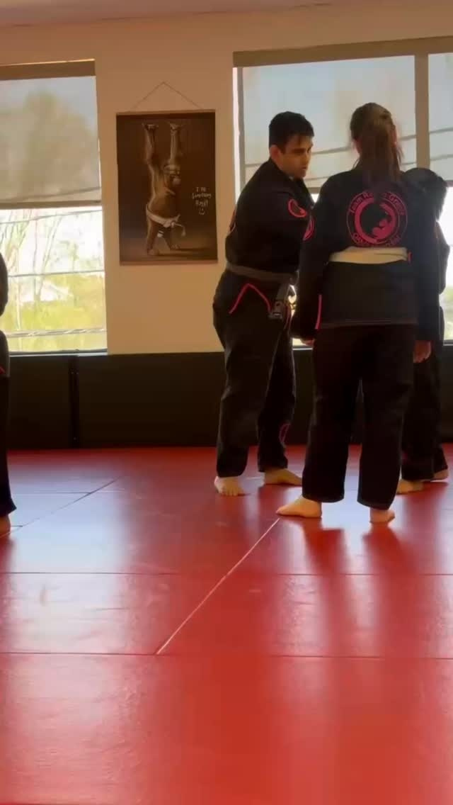 A coach in a black gi stands with a student on red mats in a bright room with windows