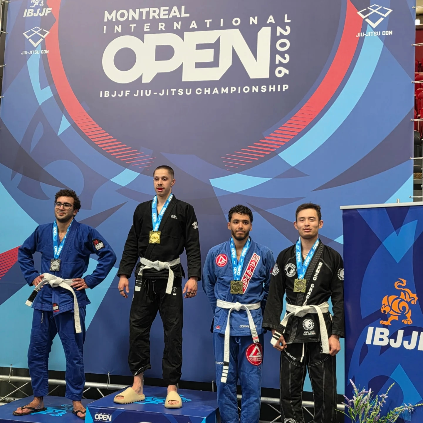 Quatre compétiteurs en kimono bleu, noir et blanc portent leur médaille sous la bannière Open 2026
