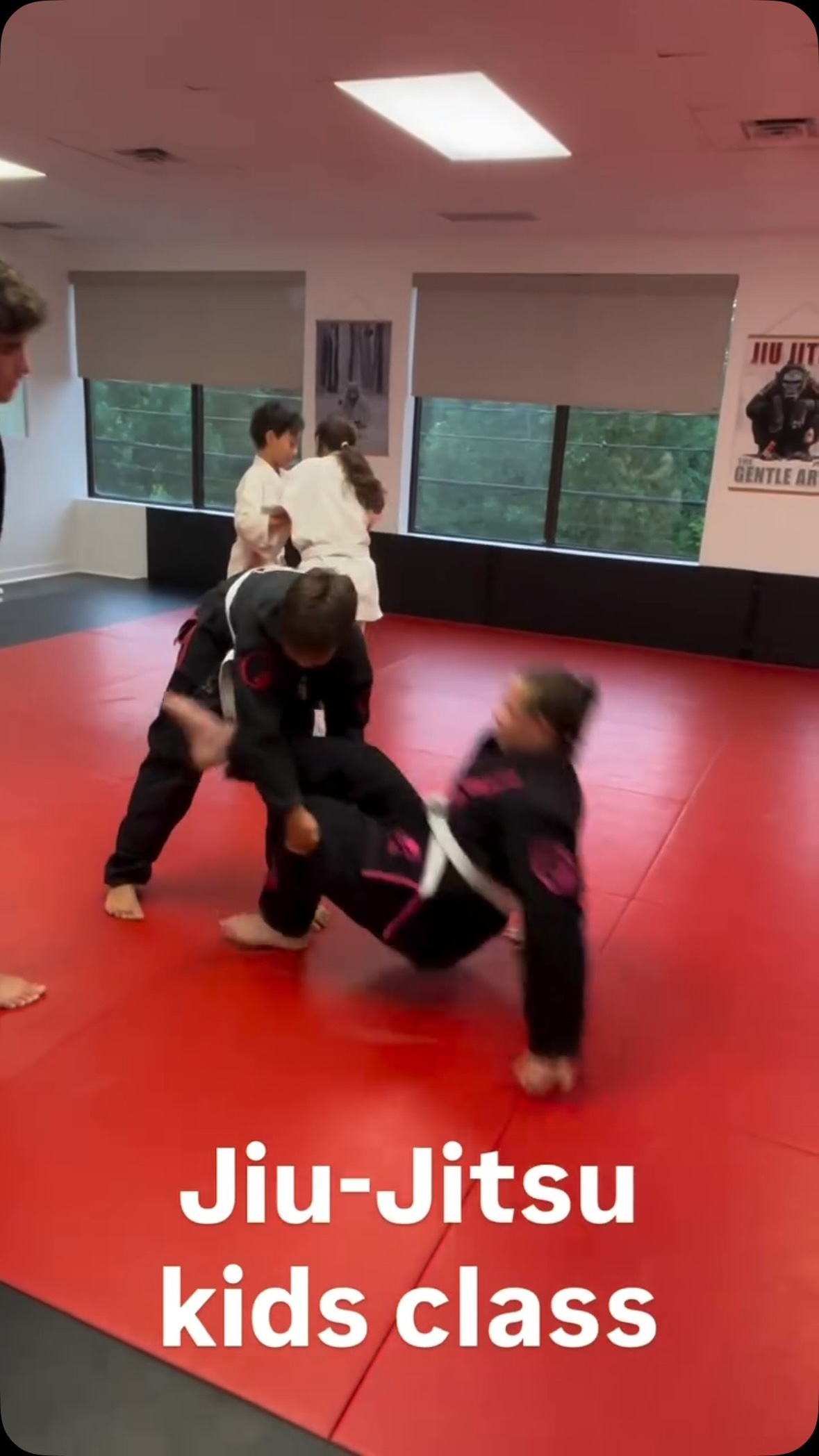 Dos alumnos jóvenes con gi negro practican un derribo sobre tatamis rojos en un salón luminoso