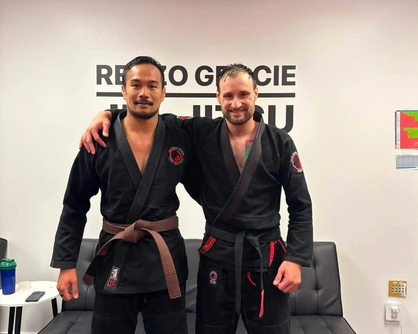Dois praticantes de kimono preto, um de faixa marrom e outro de faixa preta, abraçados