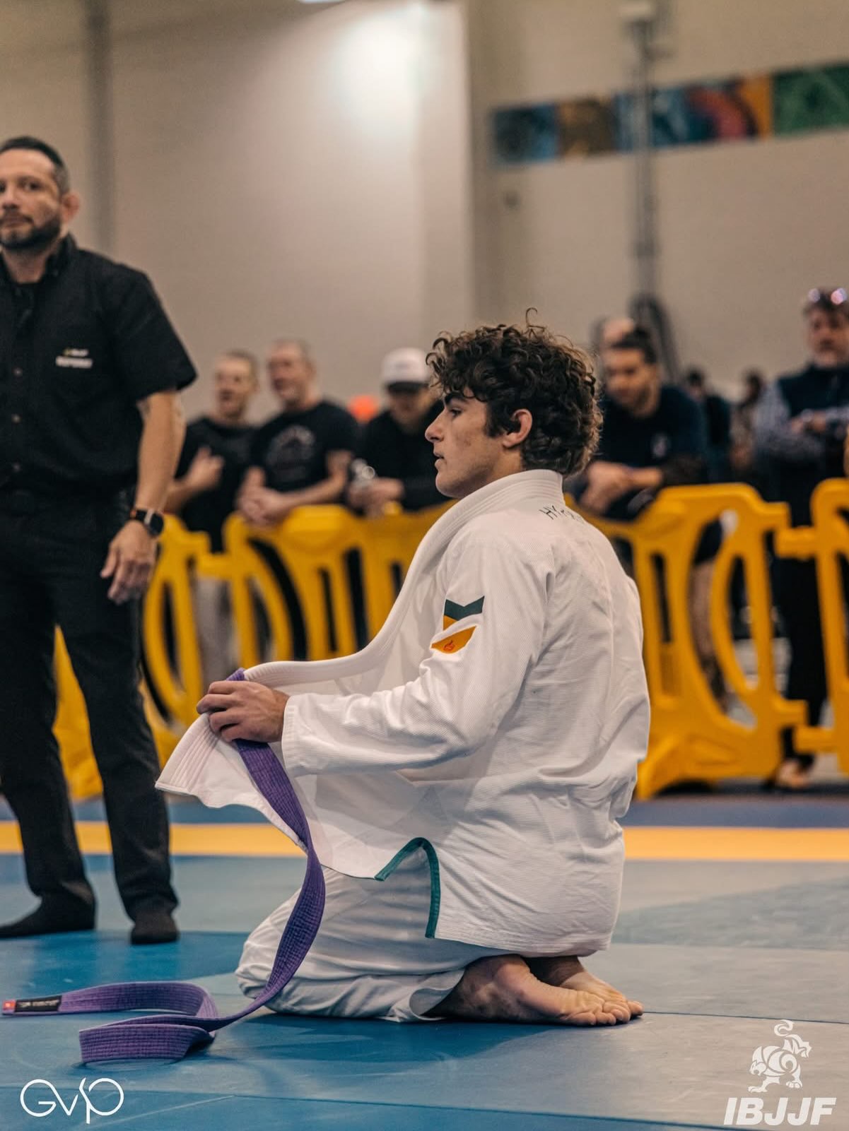 Un competidor de cinturón morado, de rodillas en el tatami, se ata el cinturón en un torneo IBJJF